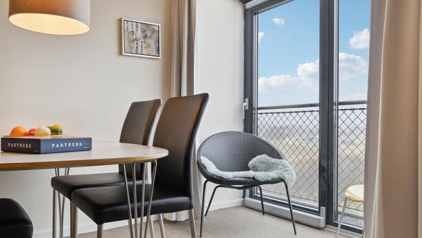 A modern hotel room with a dining table, two chairs, a bookshelf with books, a framed abstract painting, and a balcony with a view.