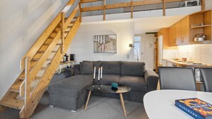 A loft-style living space with a wooden staircase, a gray sofa, a small round table with candles, and a kitchen area with wooden cabinets.