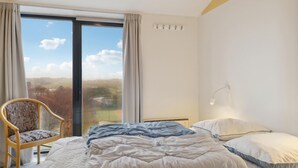 A bedroom with a bed, a chair, a window with curtains, a wall-mounted lamp, and a view of the outdoors.