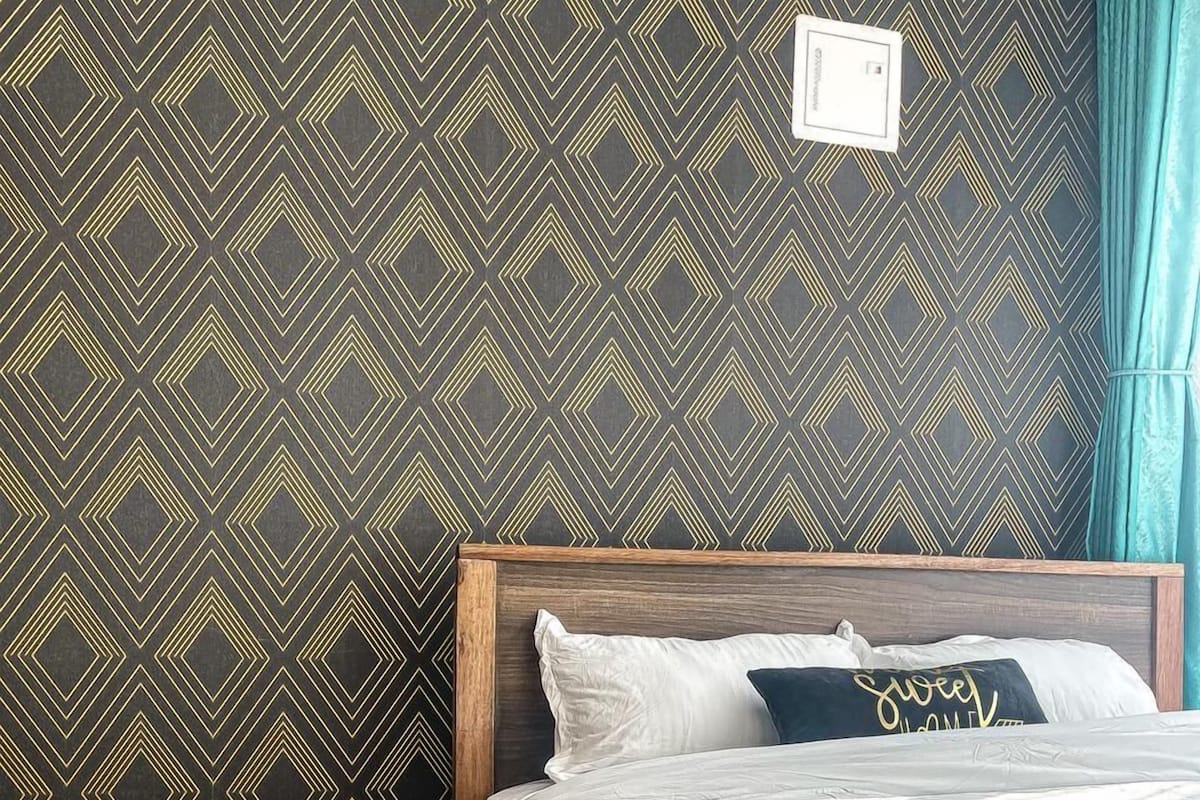 Una cama bien hecha con cabecera de madera, una pequeña mesa redonda con un control remoto y una alfombra estampada en el suelo.