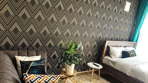 A room with a patterned wallpaper, a grey sofa with patterned cushions, a small round table with a plant, a wooden bed with white bedding, and a window with curtains. - Jiji poa (Nairobi)