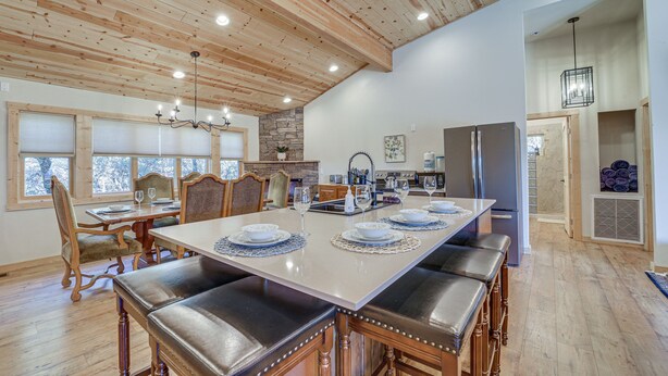 Une vaste salle à manger avec une longue table dressée pour six personnes, des poutres apparentes en bois au plafond et une cuisine en arrière-plan.