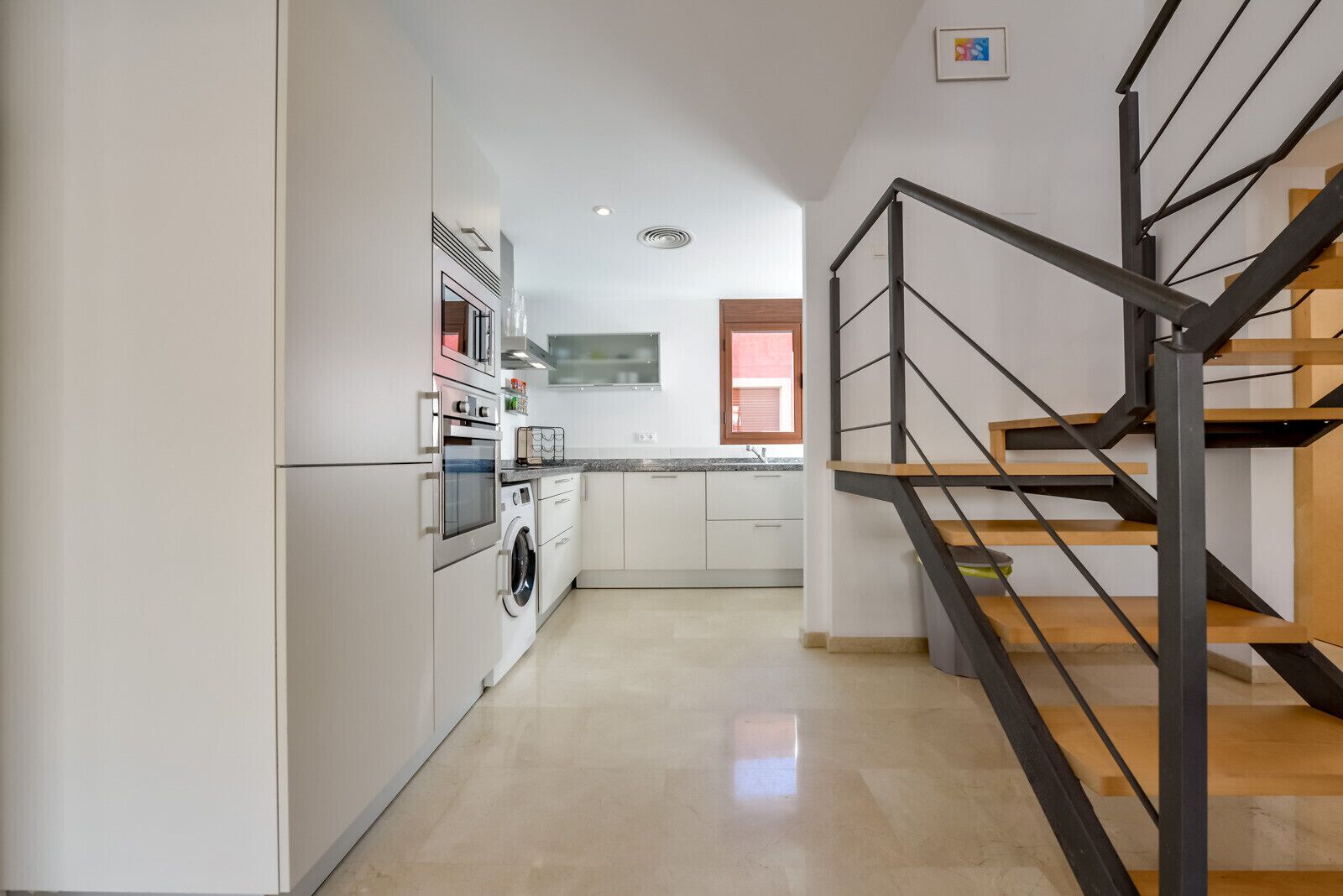 A modern kitchen with stainless steel appliances, a wooden staircase, and a view to the living area.