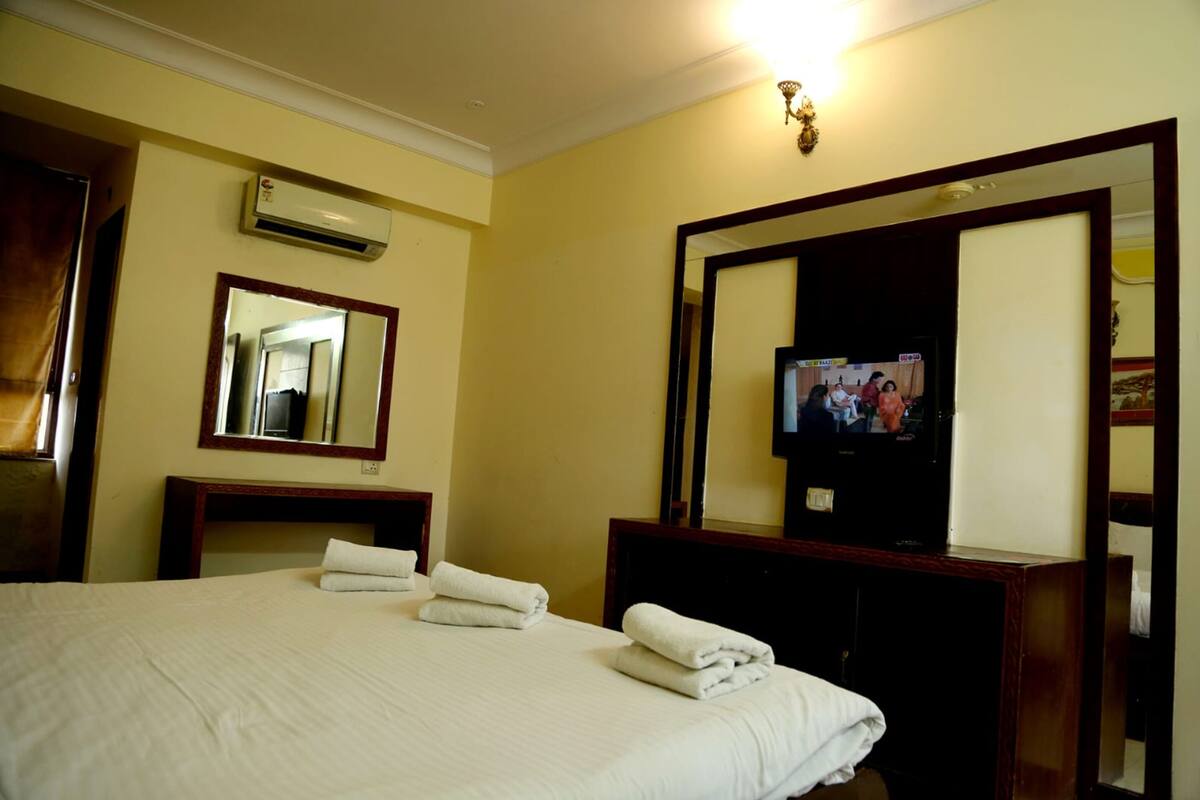 A hotel room with a bed, a TV, a mirror, and an air conditioning unit.