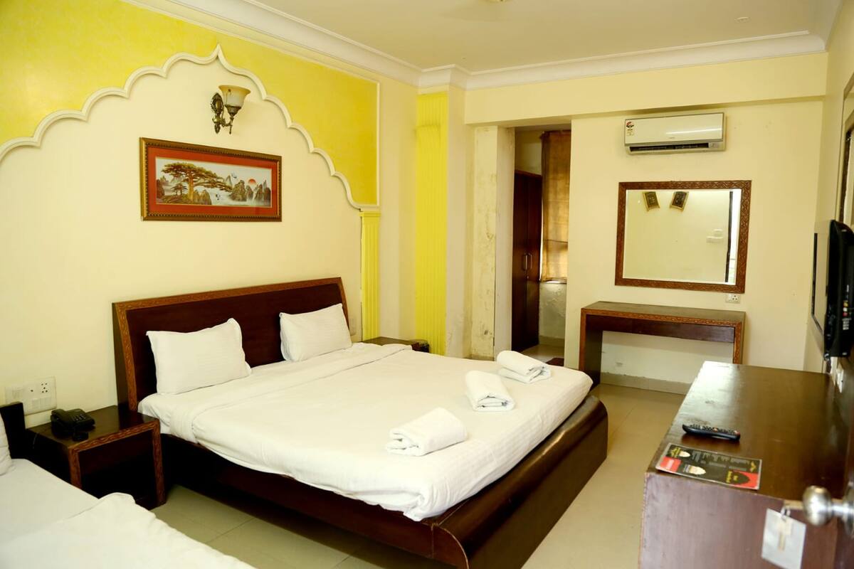 A hotel room with a bed, bedside tables, a desk, a mirror, and an air conditioning unit.