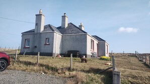 A two-story house with a stone facade, multiple chimneys, and a fenced yard with a bench and potted plants. - 27 Garenin (Isle of Lewis)