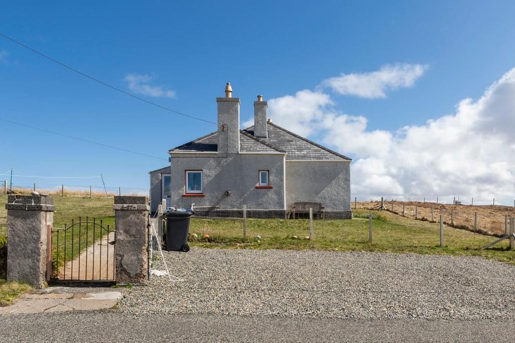A house with a gravel driveway, a metal gate, and a grassy area with a fence.