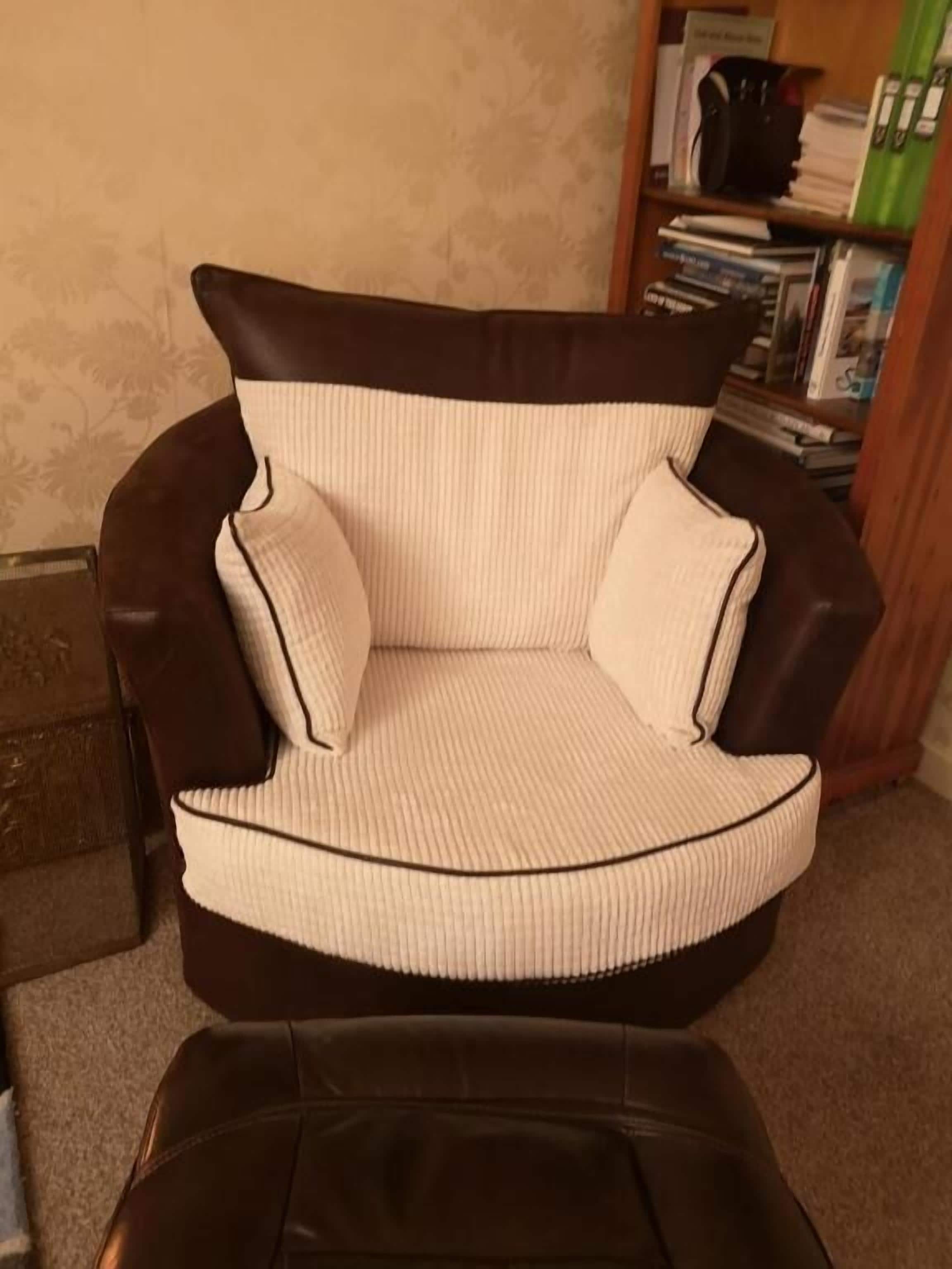 Uma poltrona de couro marrom com assento e encosto acolchoados em branco, em um quarto com uma estante de livros ao fundo.