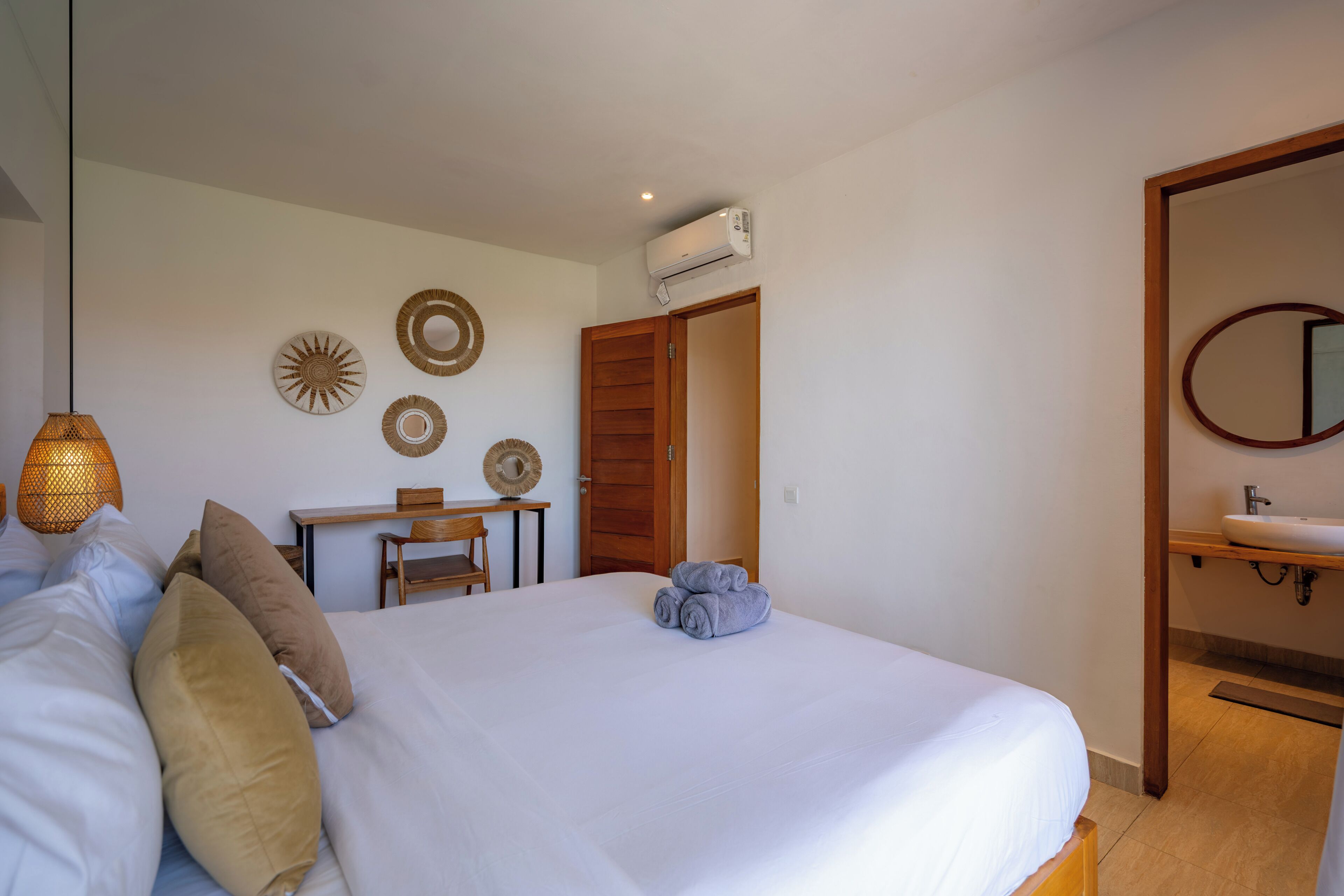 Una cama bien hecha con almohadas, una mesita con una silla, un aire acondicionado instalado en la pared y un baño con lavabo y espejo.