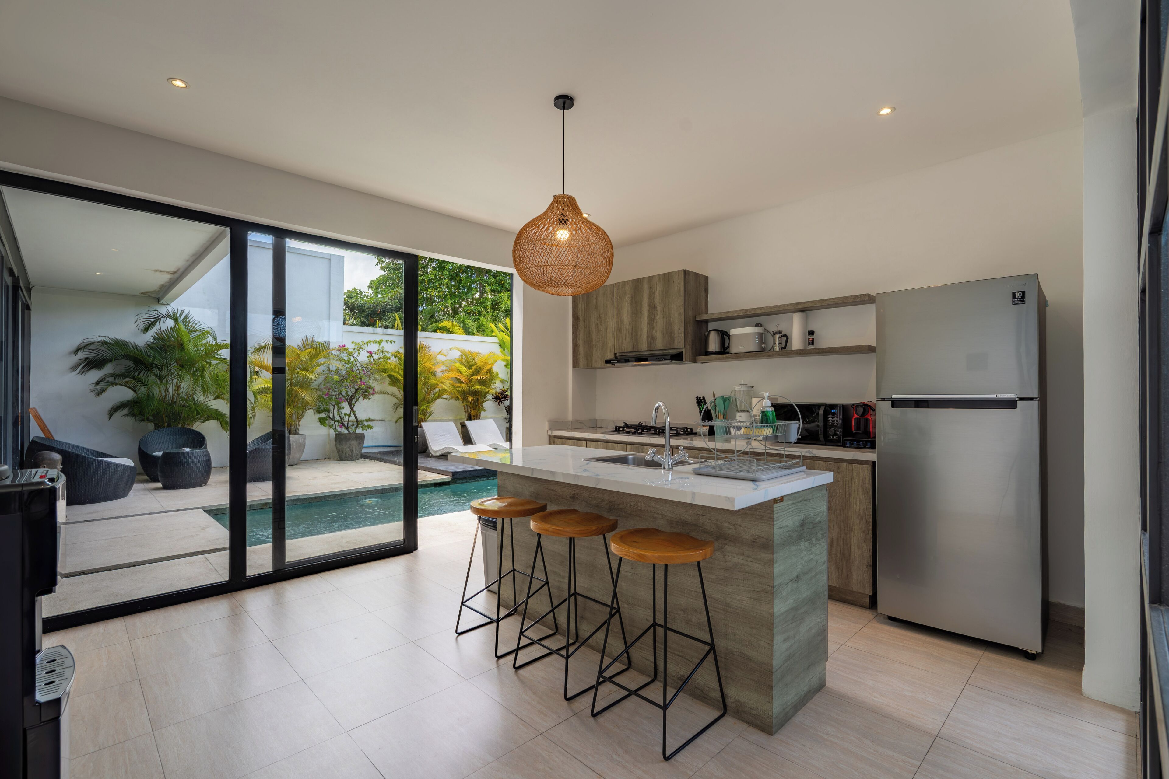 A modern kitchen with a large island, bar stools, a refrigerator, and a view of a swimming pool.