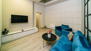 A modern living room with a flat-screen TV mounted on the wall, a sofa set, a coffee table with a vase of flowers, and a window providing natural light.