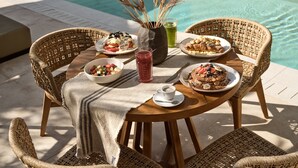Una mesa de comedor con sillas de mimbre, una piscina al fondo y diferentes platos y bebidas.