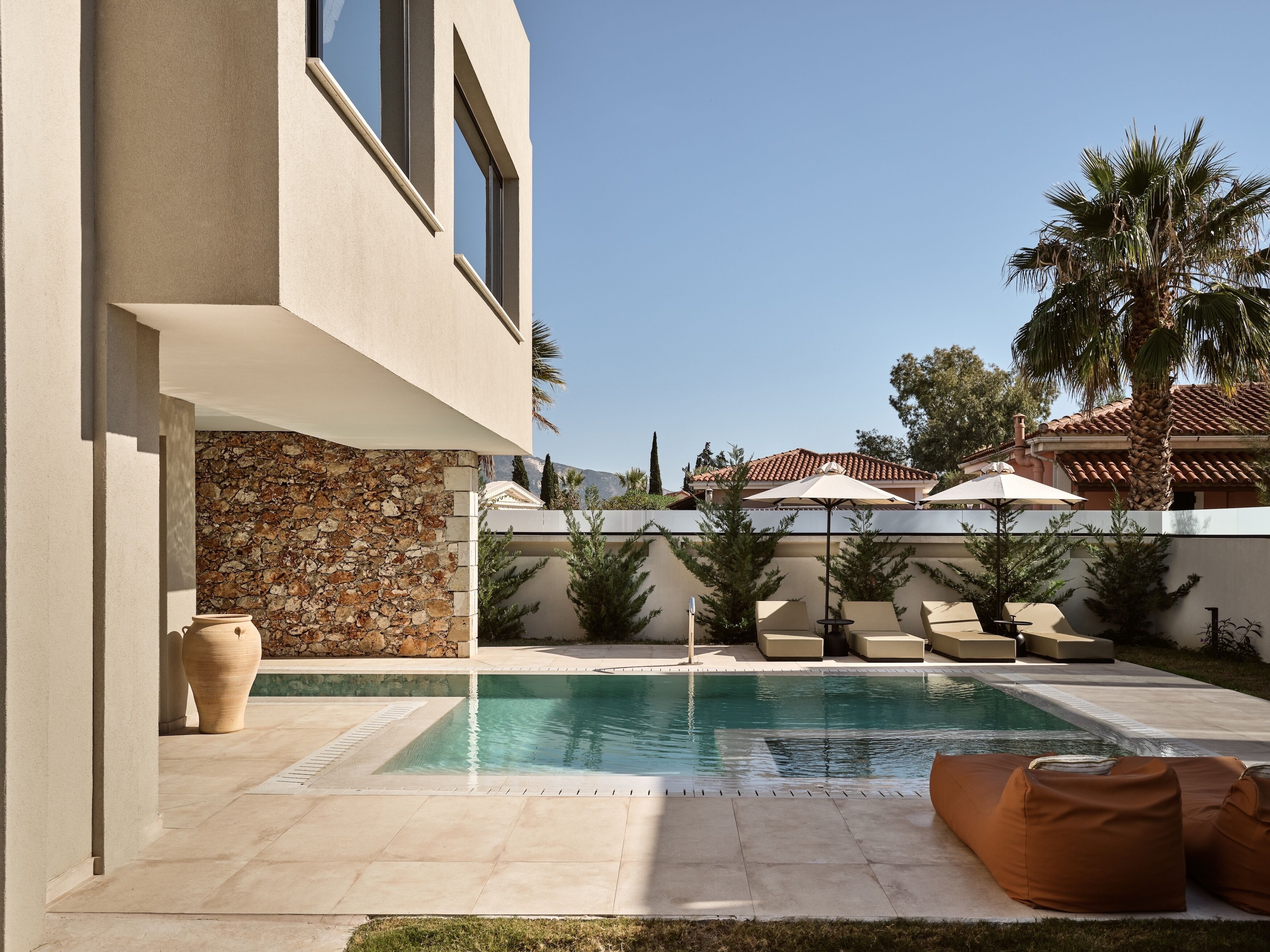 A modern pool area with lounge chairs and umbrellas, a stone wall, and a view of a mountain.