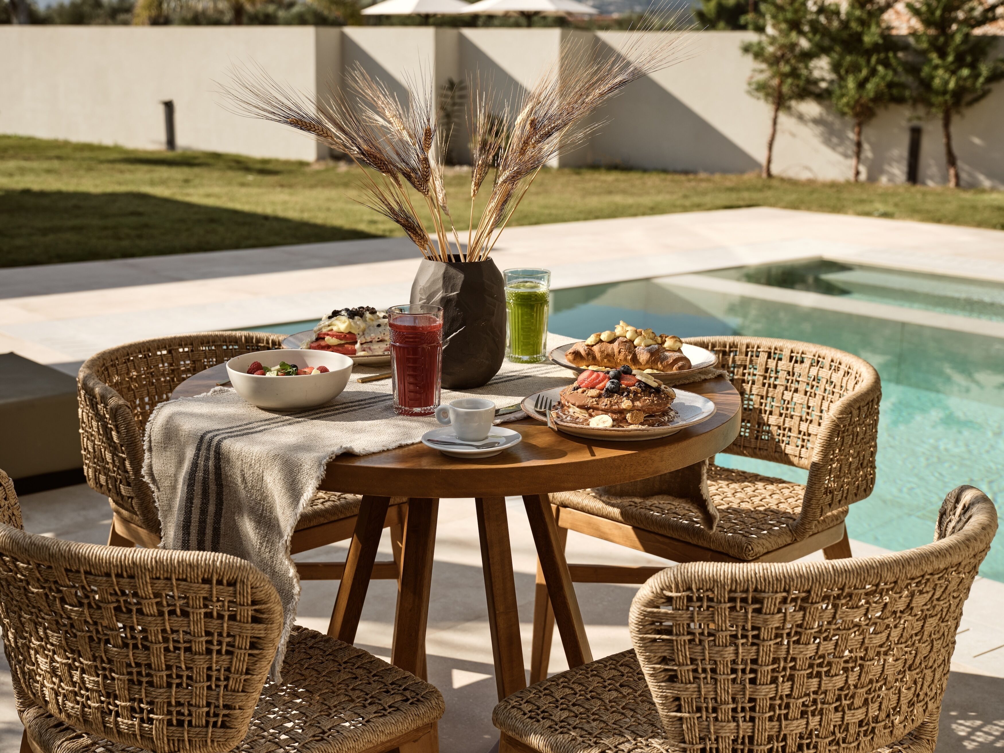Una mesa de madera redonda con sillas de mimbre, un tazón de fruta, un vaso de té helado y un plato de panqueques junto a una piscina.