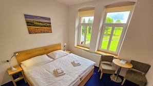 A bedroom with a wooden bed, white linens, a bedside lamp, a small table with a chair, and a window with a view of greenery.