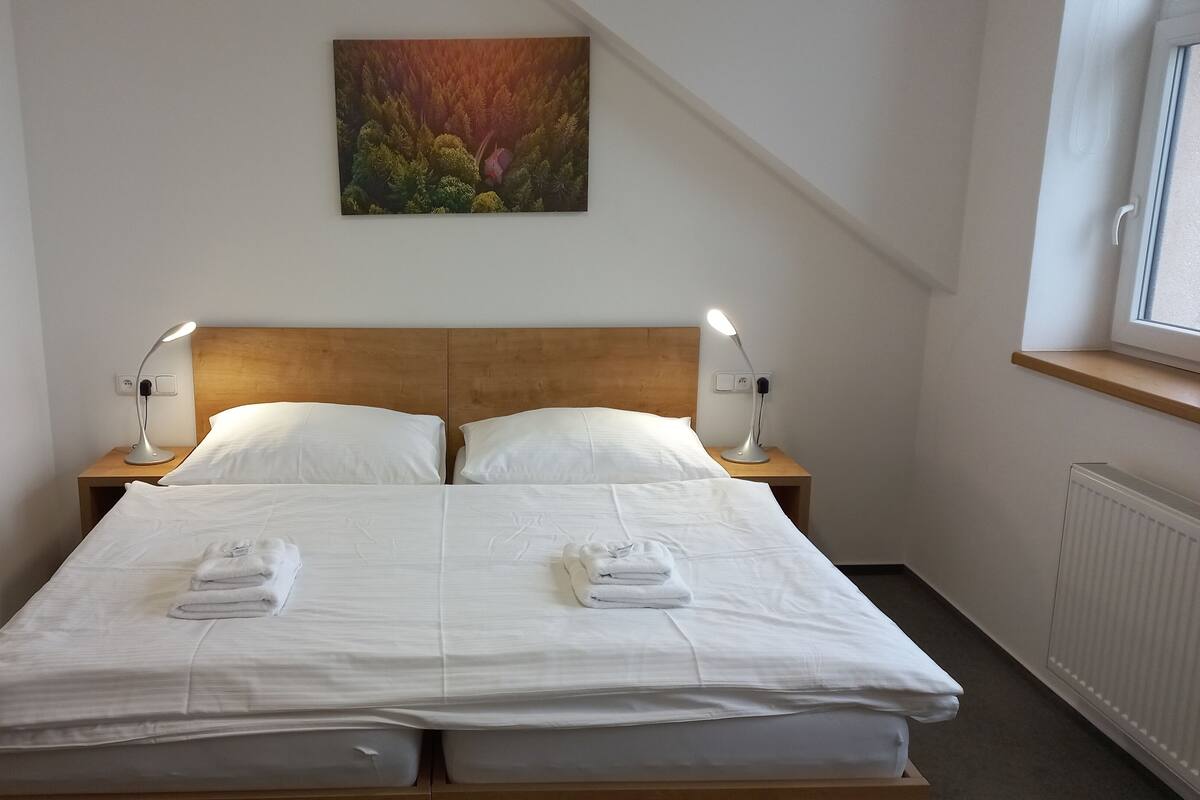 Un lit double avec du linge de lit blanc et deux tables de chevet avec des lampes, un tableau au mur et une fenêtre avec une étagère en bois.