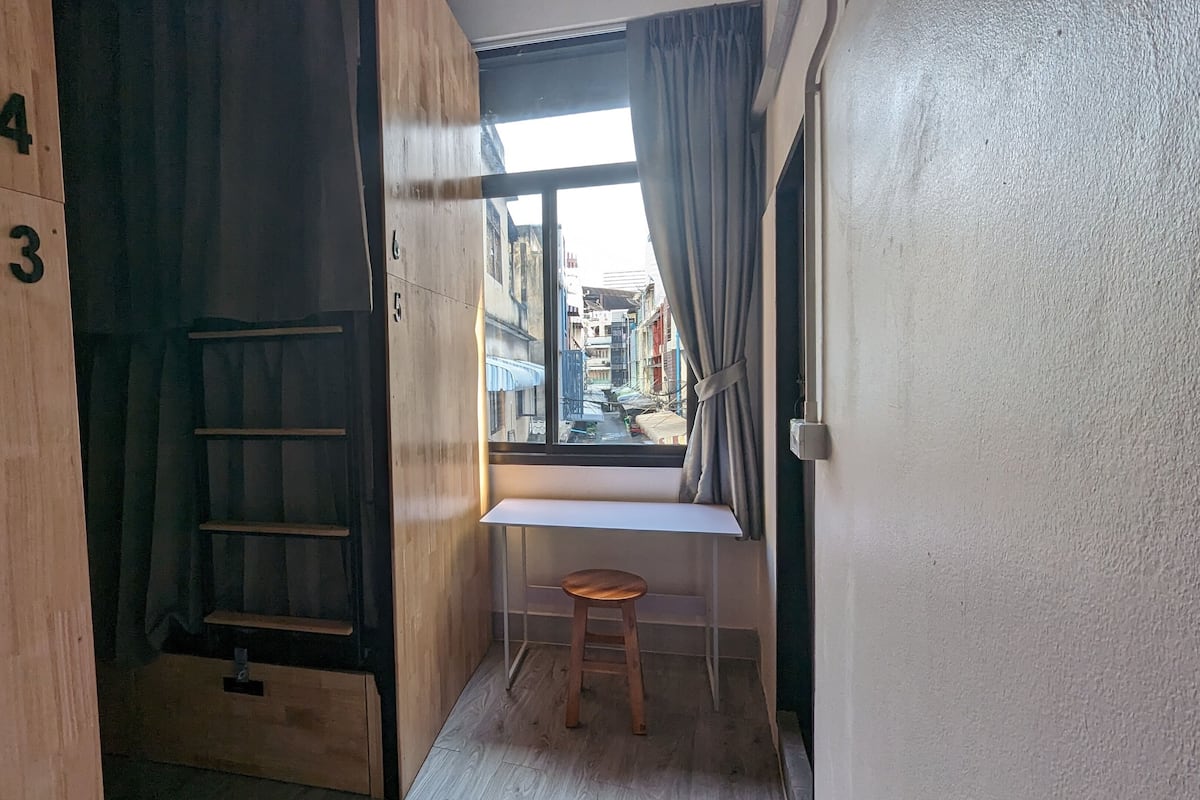 Une petite pièce étroite, avec une armoire en bois, une  échelle, une fenêtre avec des rideaux et un petit tabouret.