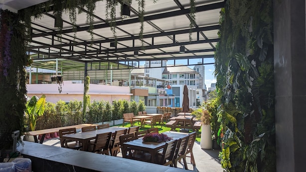 Un área de comedor al aire libre con mesas y sillas de madera, rodeada de abundante vegetación y un techo de cristal.