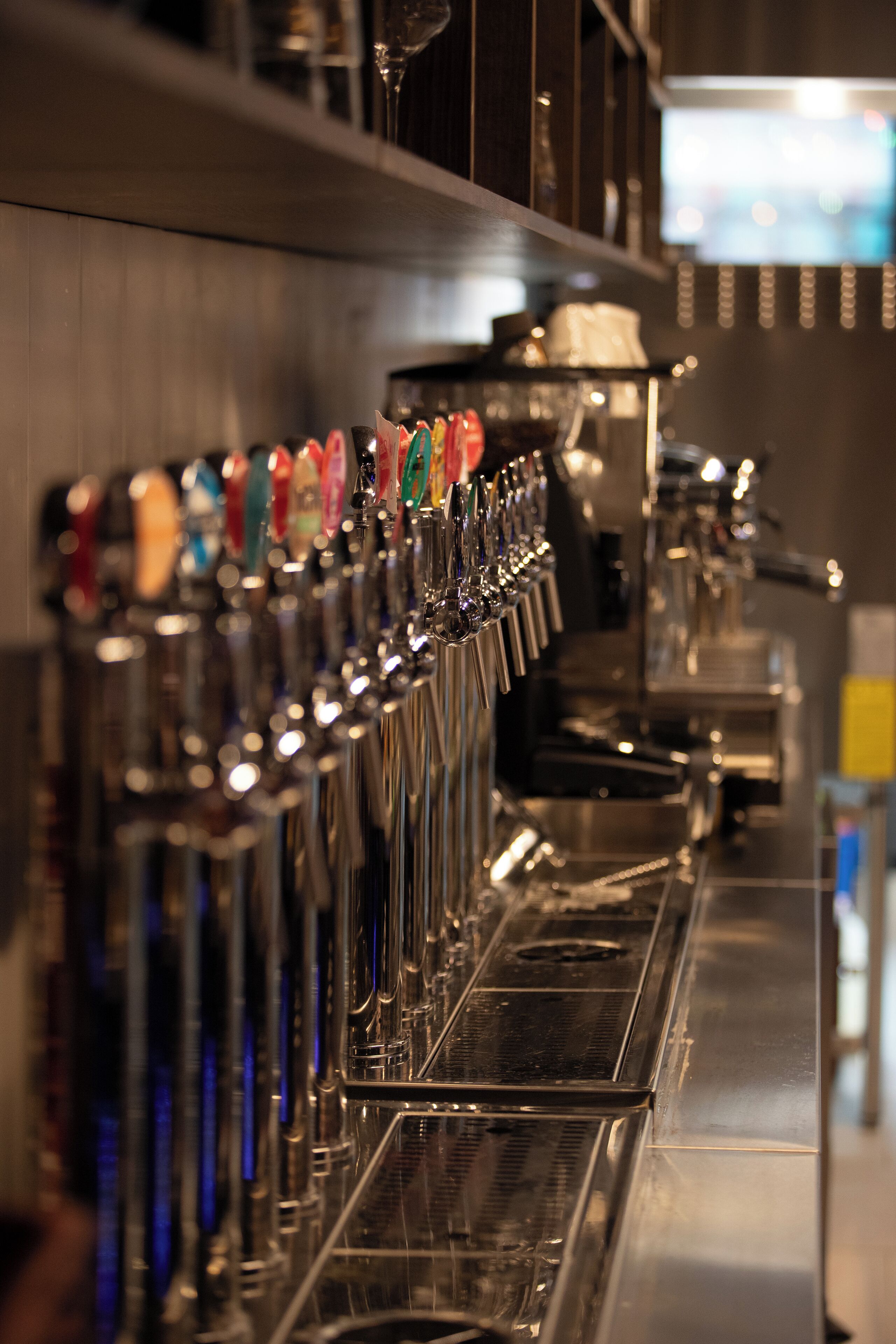 Une rangée de robinets à bière aux poignées colorées, un appareil à café et un écran de télévision en arrière-plan.