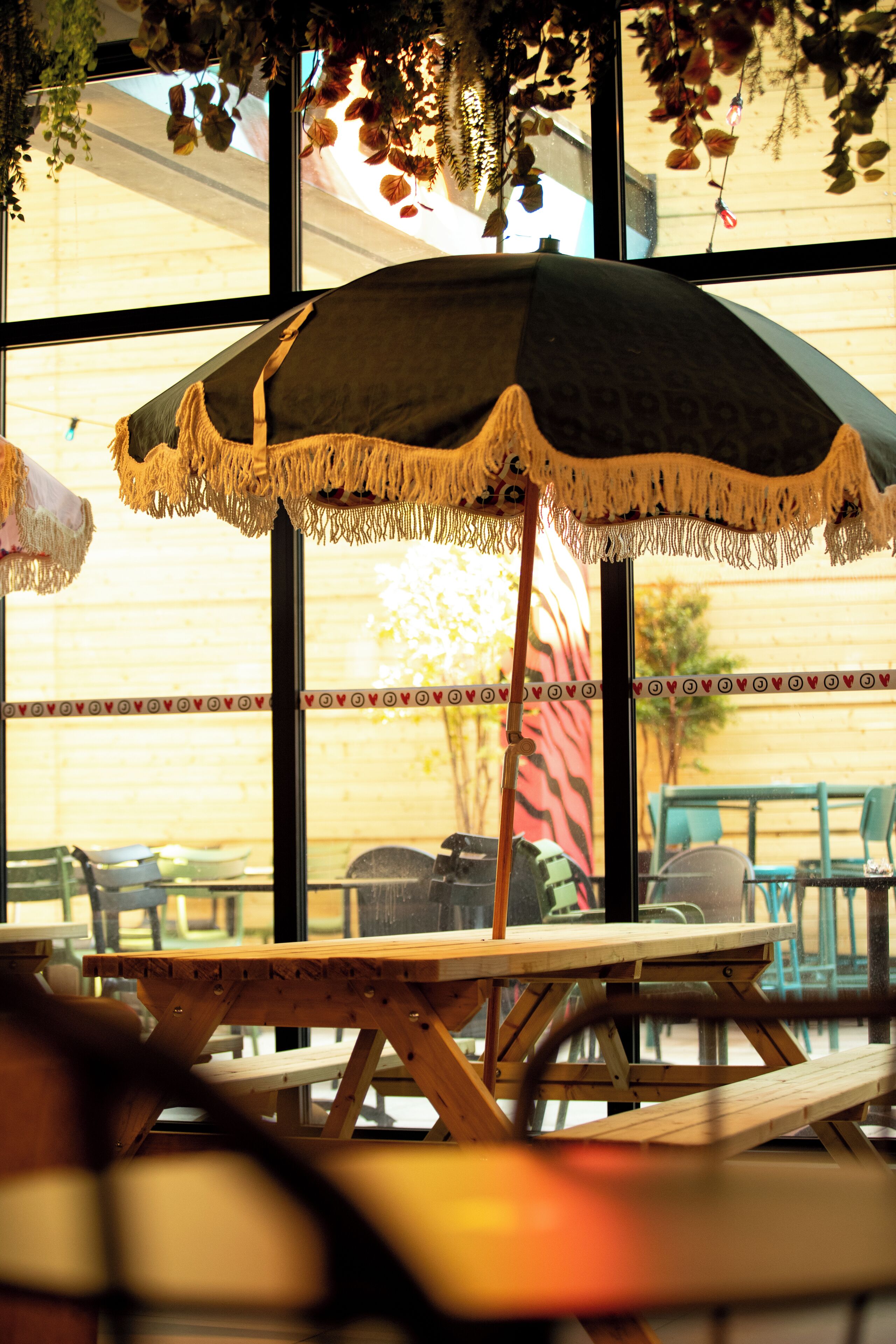 A wooden table with a green umbrella, a wooden bench, and a view of outdoor seating through large windows.