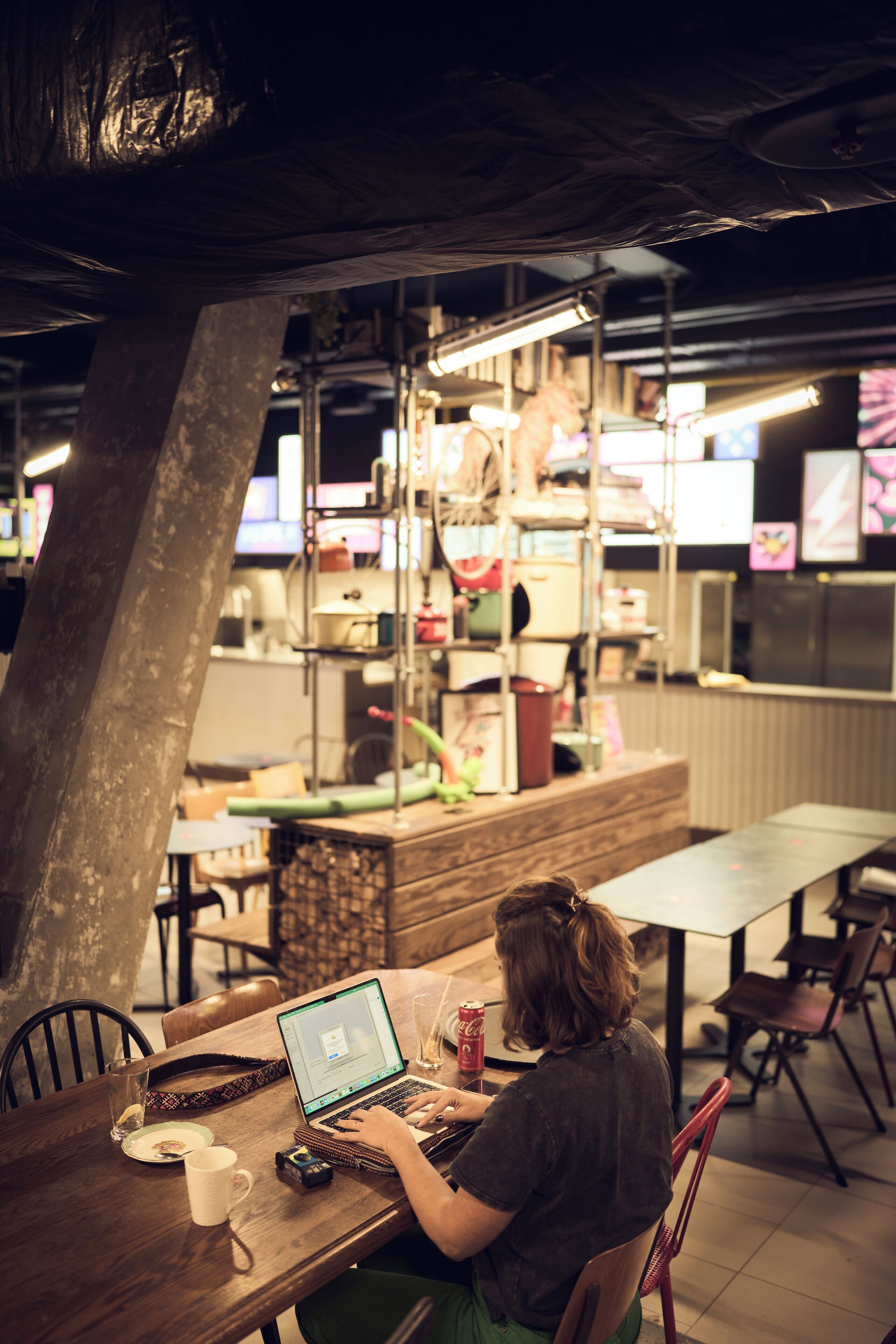 Una persona trabajando en un ordenador portátil sobre una mesa de madera en una cafetería, con una barra y estanterías al fondo.