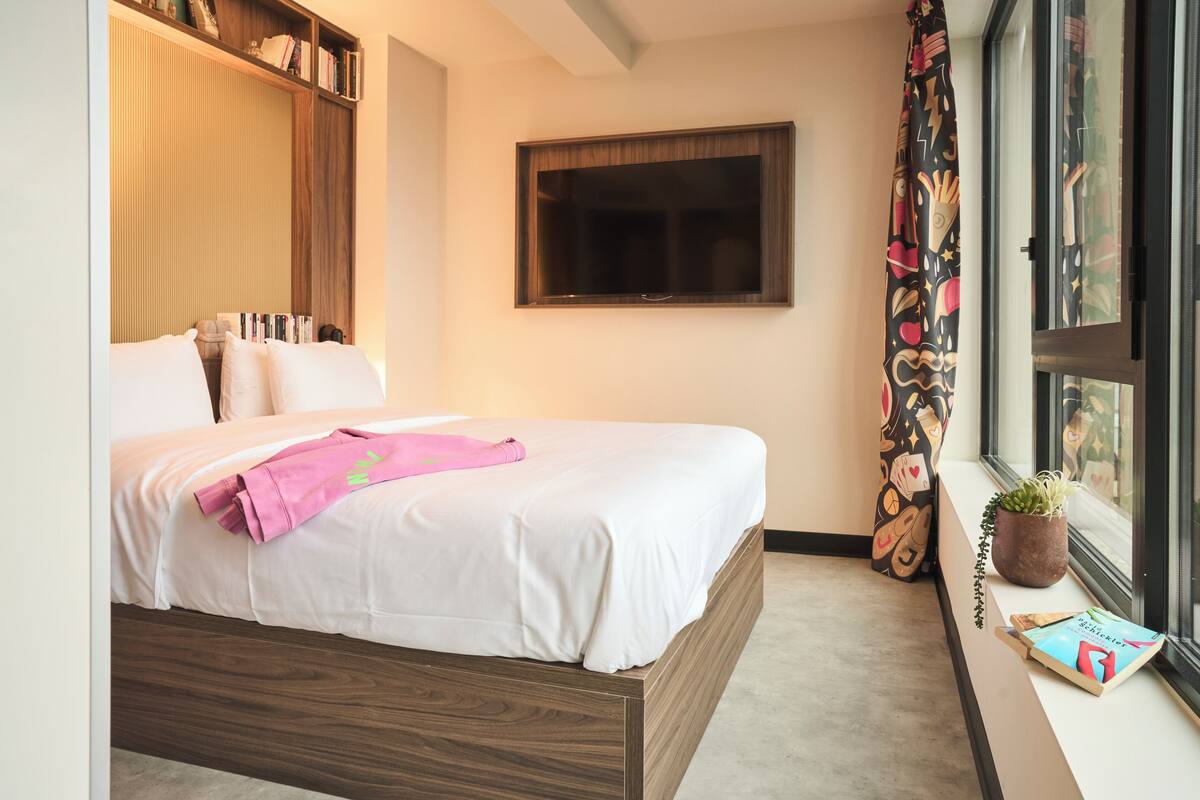 Een keurig opgemaakt bed met een witte dekbedovertrek en roze handdoeken, een houten hoofdbord, een groot raam met uitzicht en een kleine potplant op de vensterbank.