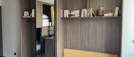 Una habitación compacta con armario empotrado, espejo, una pequeña zona de estar y una estantería con libros y objetos decorativos.
