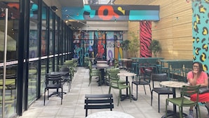 An outdoor cafe with tables and chairs, a large mural on the wall, and a staircase leading to an upper level.