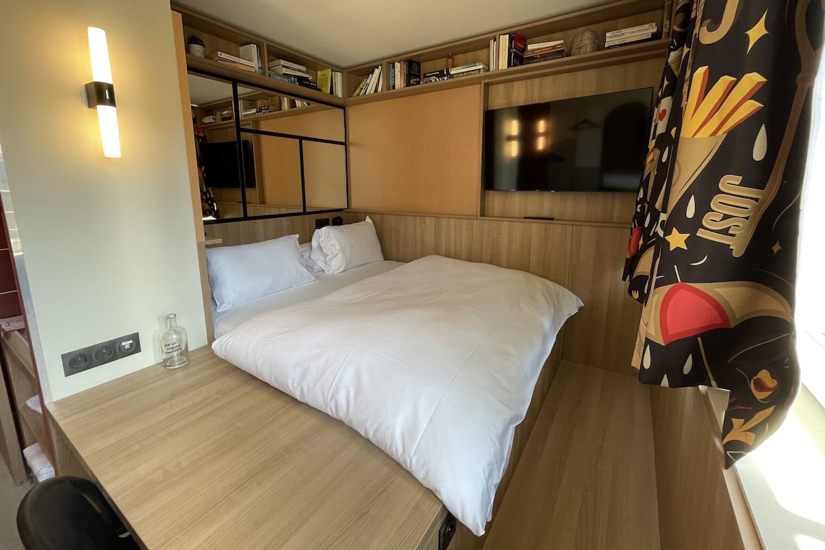 Een knus slaapkamer met een bed, een televisie, een boekenplank en een raam met gordijnen.