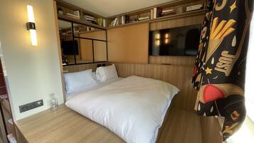 Un dormitorio pequeño con cama, televisor, estantería y ventana con cortinas.
