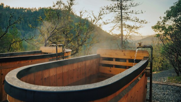 Een houten jacuzzi met een gebogen rand, omgeven door een houten terras, met op de achtergrond bomen en heuvels.