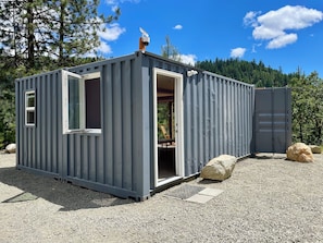A compact, grey metal building with a door open, revealing an interior with a chair and table. - Off-grid container home on 6 private acres, outdoor soaking tubs and plunge pool (Rogue River)