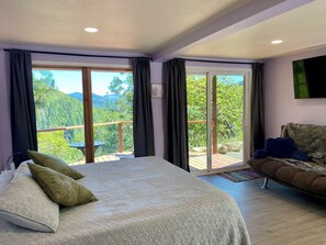 A bedroom with a bed, pillows, a sofa, a TV, and large sliding glass doors leading to a balcony with a view of greenery and mountains.
