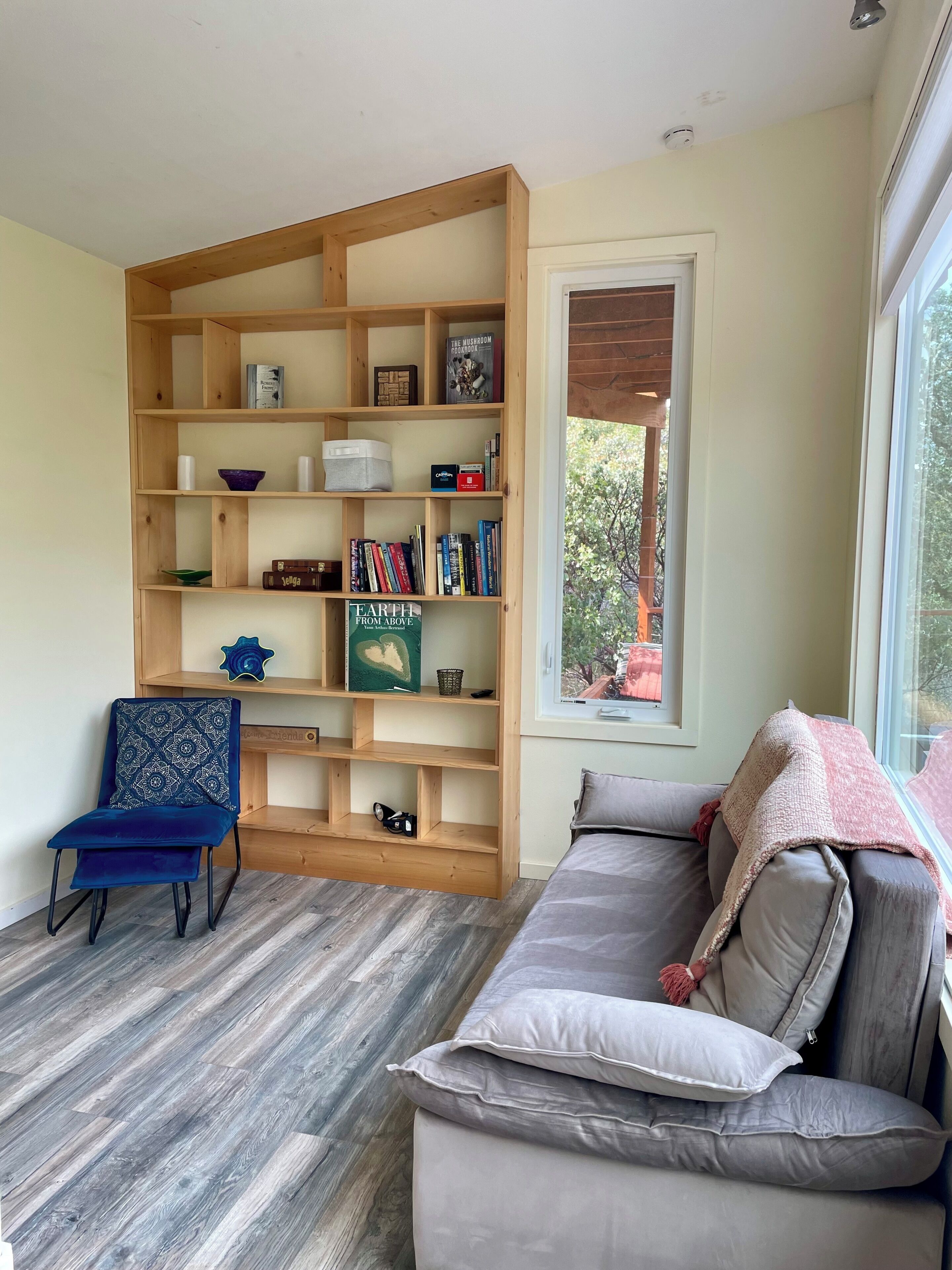 A living room with a wooden bookshelf, a blue chair, a grey sofa, and a window with a view of trees.