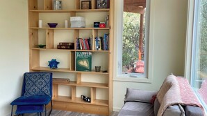 Een woonkamer met een houten boekenkast, een blauwe stoel, een grijze bank en een raam met uitzicht op bomen.