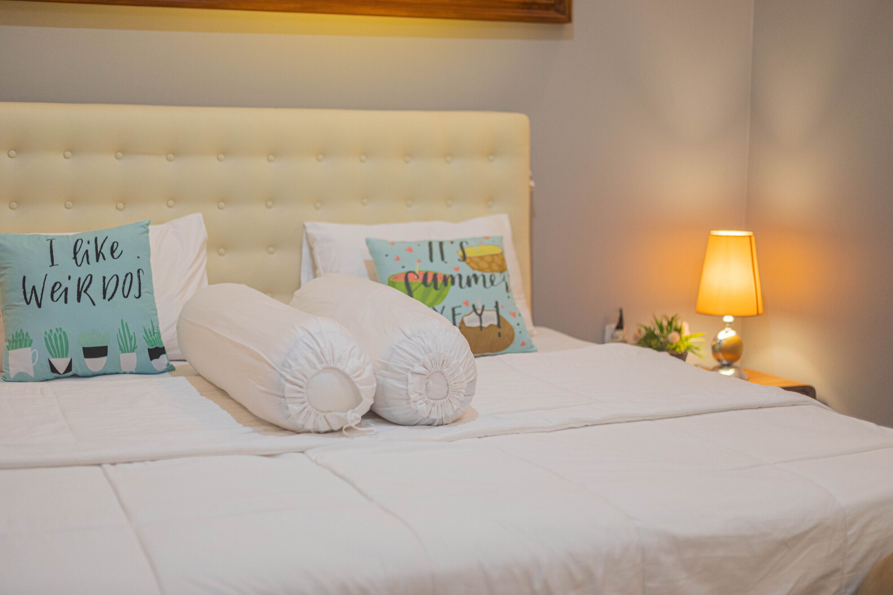 A neatly made bed with white bedding, two decorative pillows with text, and a bedside lamp.