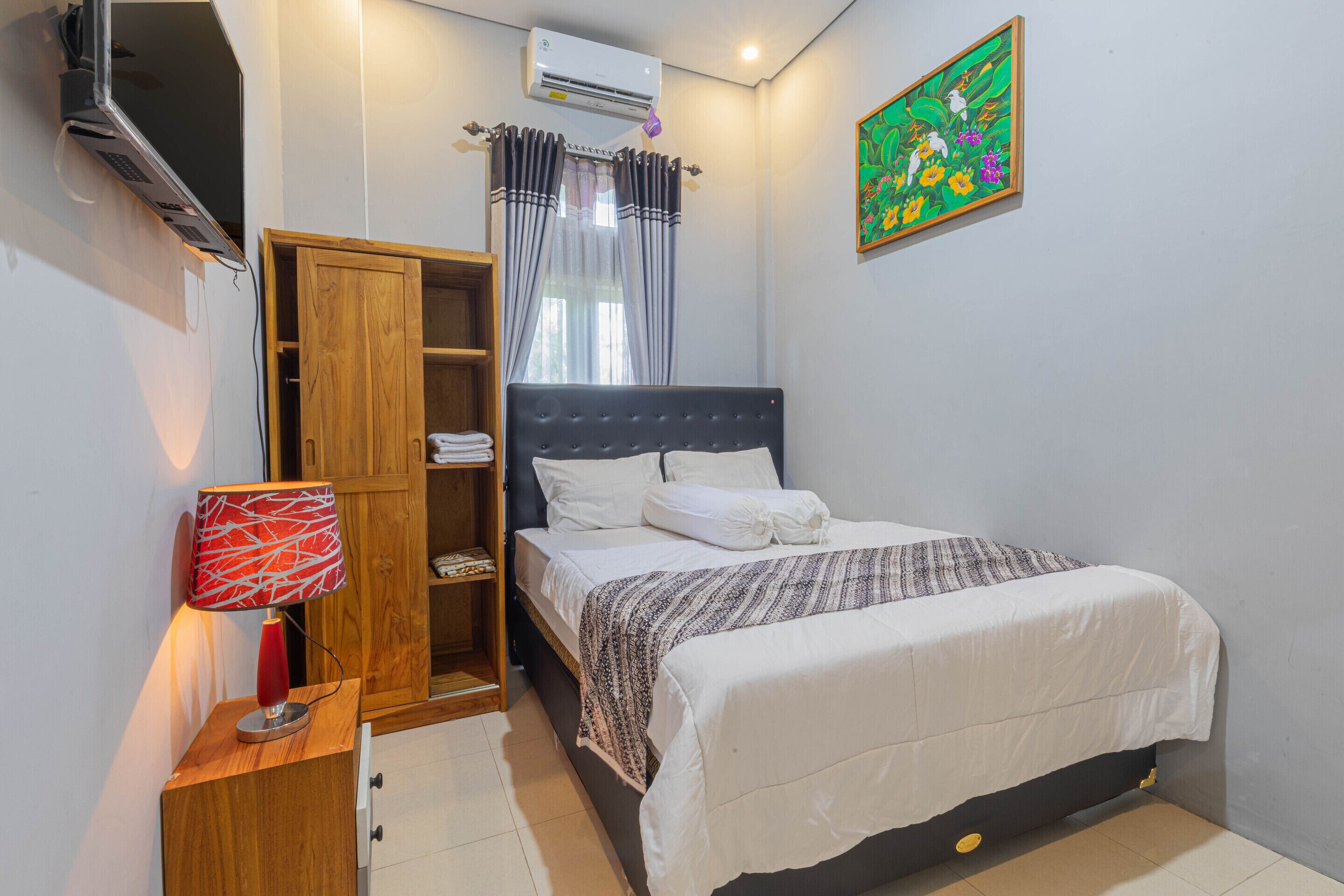 Un dormitorio con cama, armario, escritorio con lámpara, aire acondicionado y un cuadro en la pared.