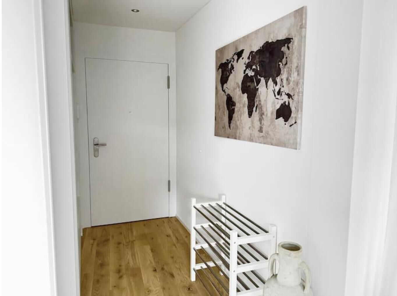 A hallway with a wooden floor, a white door, a white rack with shelves, and a world map on the wall.