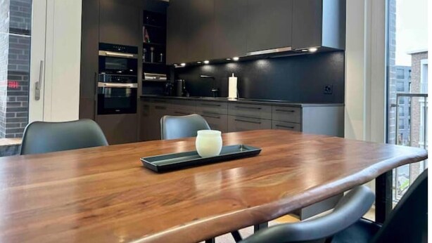 A modern kitchen with dark cabinetry, a wooden dining table, and a view of the city through large windows. - Elegante Unterkunft in Stadtnähe (Adliswil)