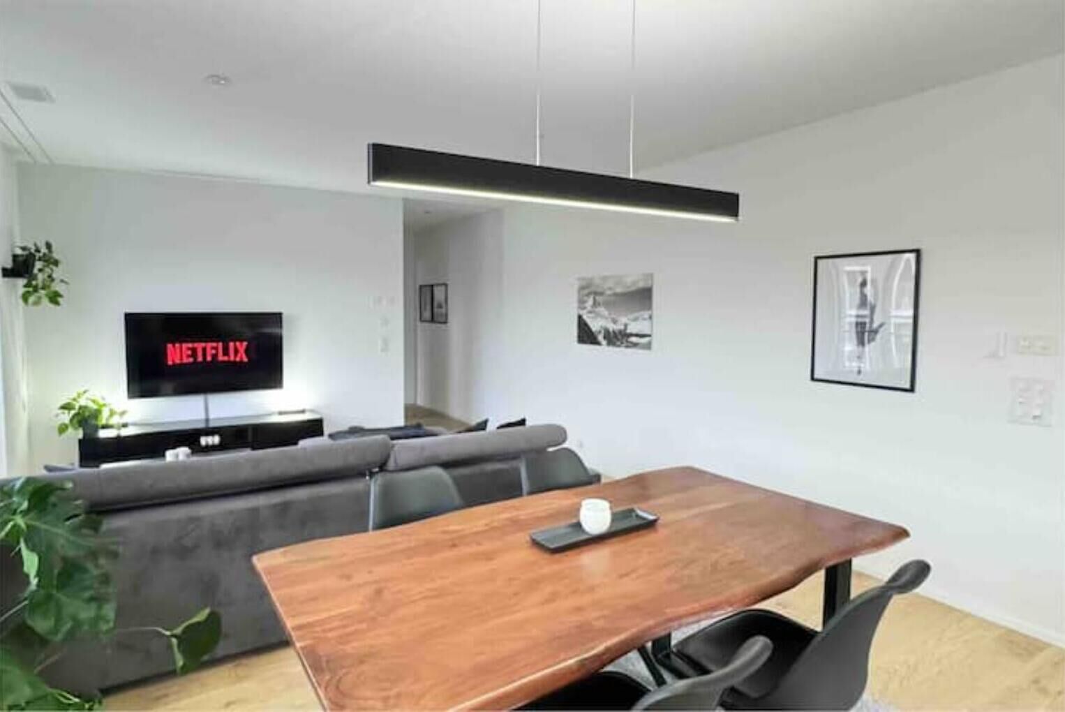 Una sala de estar moderna con una mesa de comedor de madera, un sofá gris y un televisor de pantalla plana mostrando 'NETFLIX'.