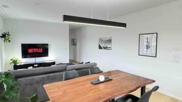 Una sala de estar moderna con una mesa de comedor de madera, un sofá gris y un televisor de pantalla plana mostrando 'NETFLIX'.
