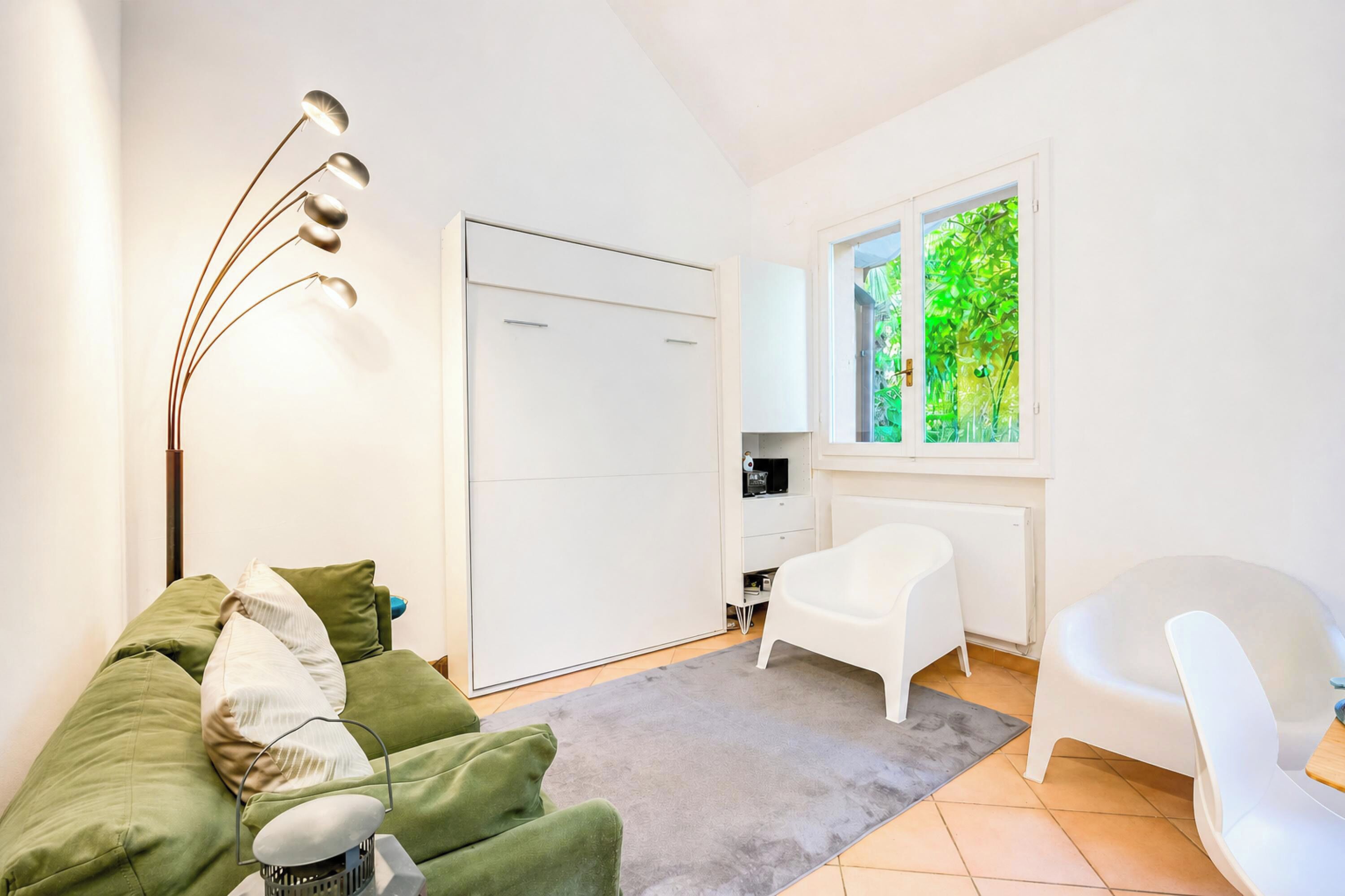 A room with a green sofa, a white chair, a floor lamp, a wardrobe, and a window with a view of greenery.