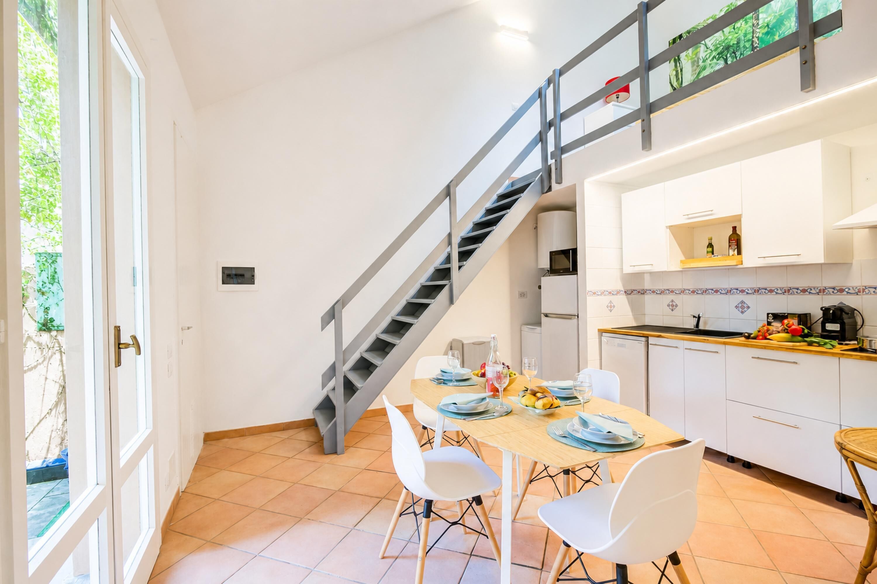 A modern kitchen with white cabinets, a dining area with a wooden table and white chairs, and a staircase with a metal railing.