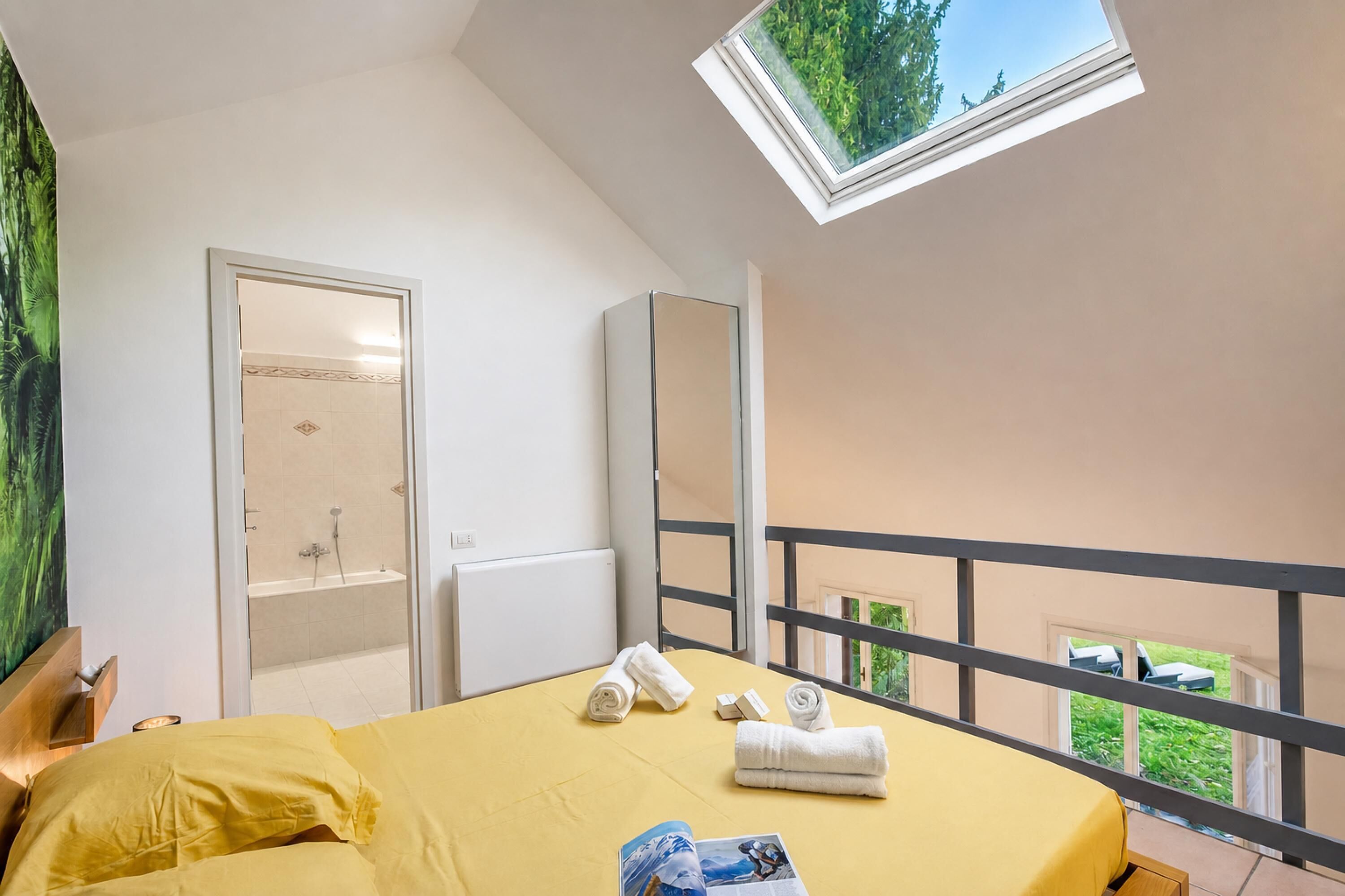 Et soveværelse med ovenlys, en seng med gul bemærkning, et badeværelse med bruser og en balkon med gelænder.