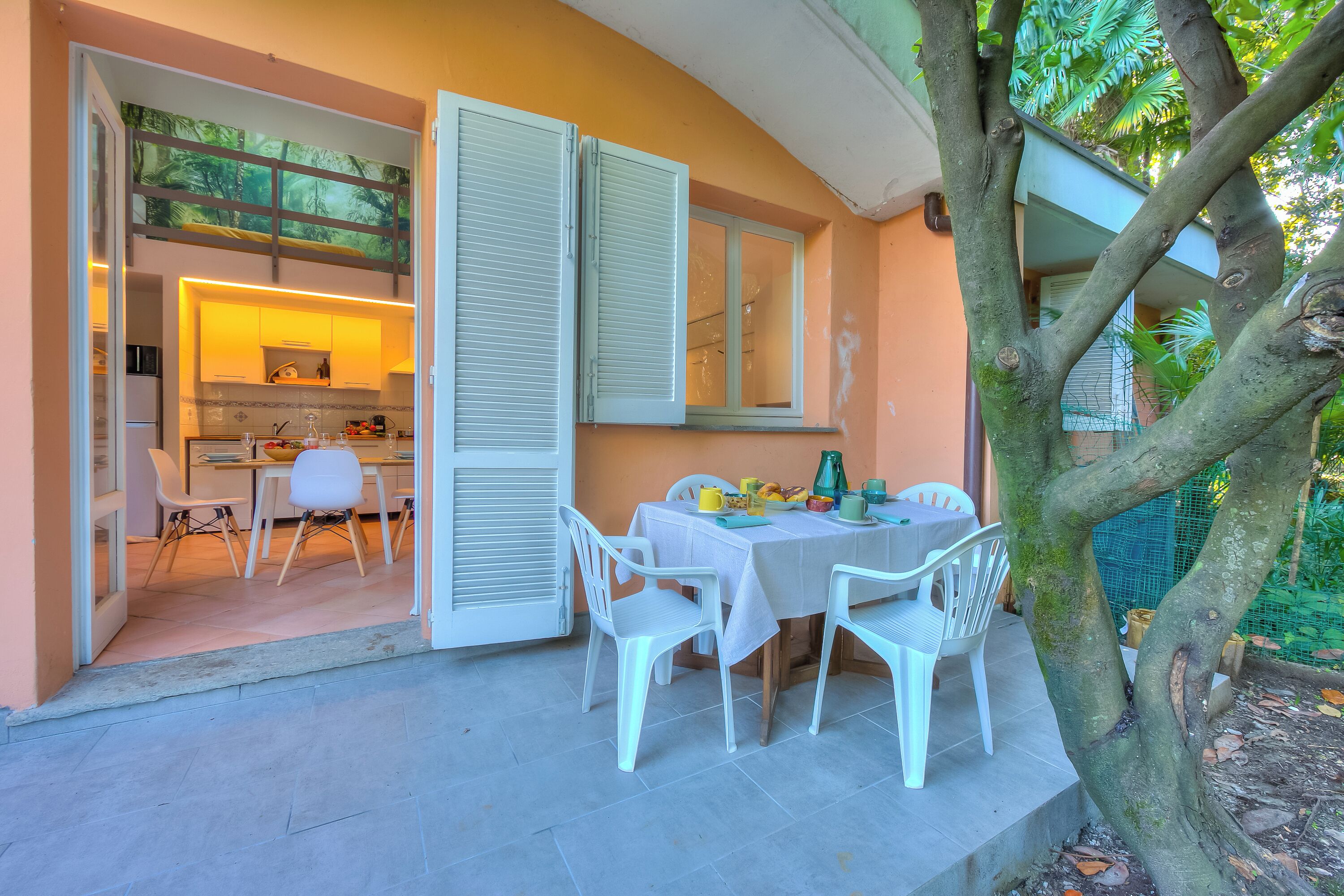 An open dining area with a table and chairs, a kitchen in the background, and a tree in the foreground.