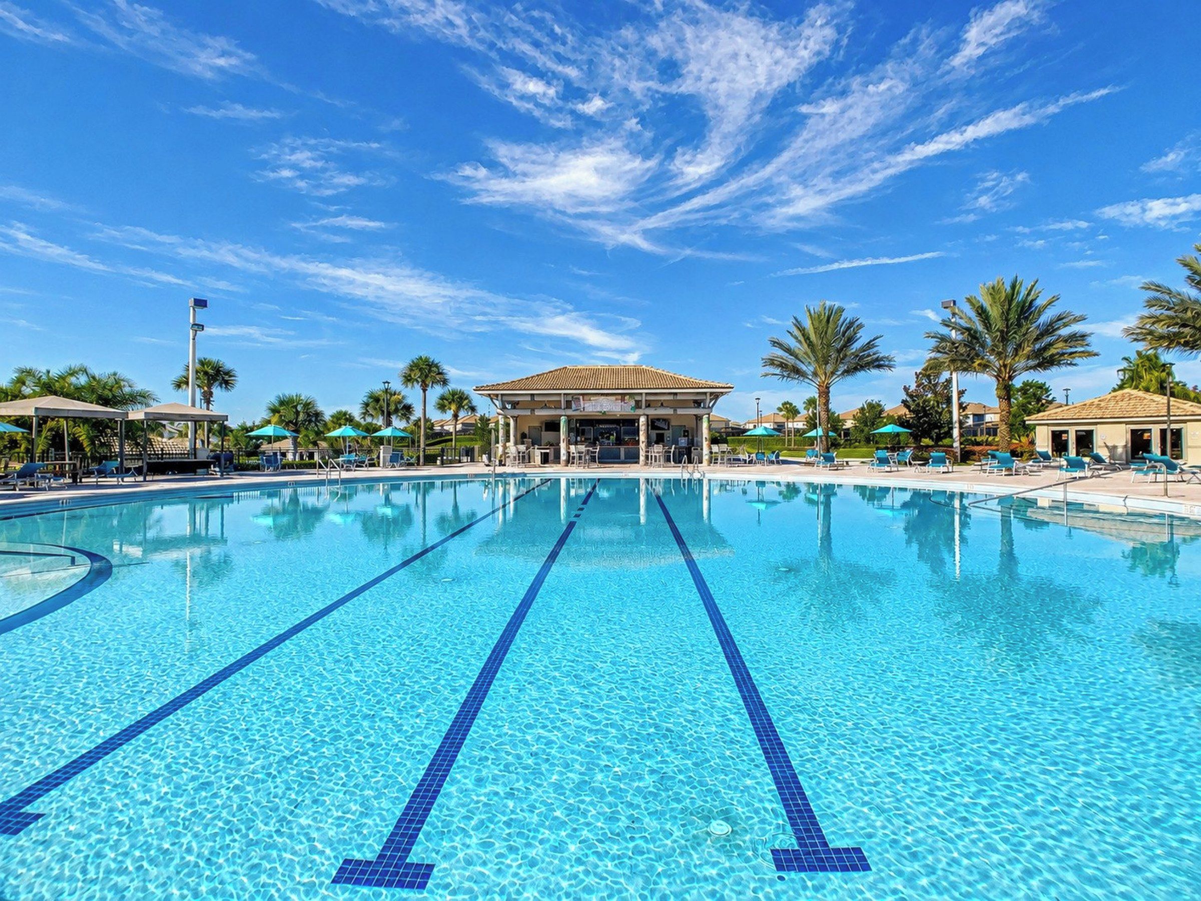 A clear blue swimming pool with lounge chairs and umbrellas, surrounded by palm trees and a pavilion.
