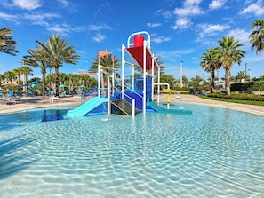 A clear pool with a water slide and a red structure, surrounded by palm trees and lounge chairs.