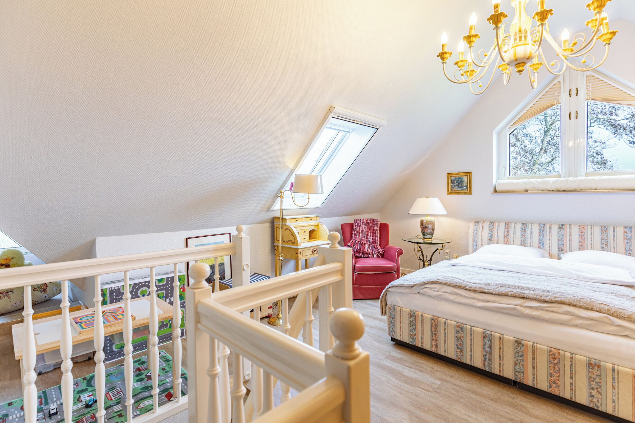 A bedroom with a sloped ceiling, a bed, a window, a chandelier, and a toy train set.