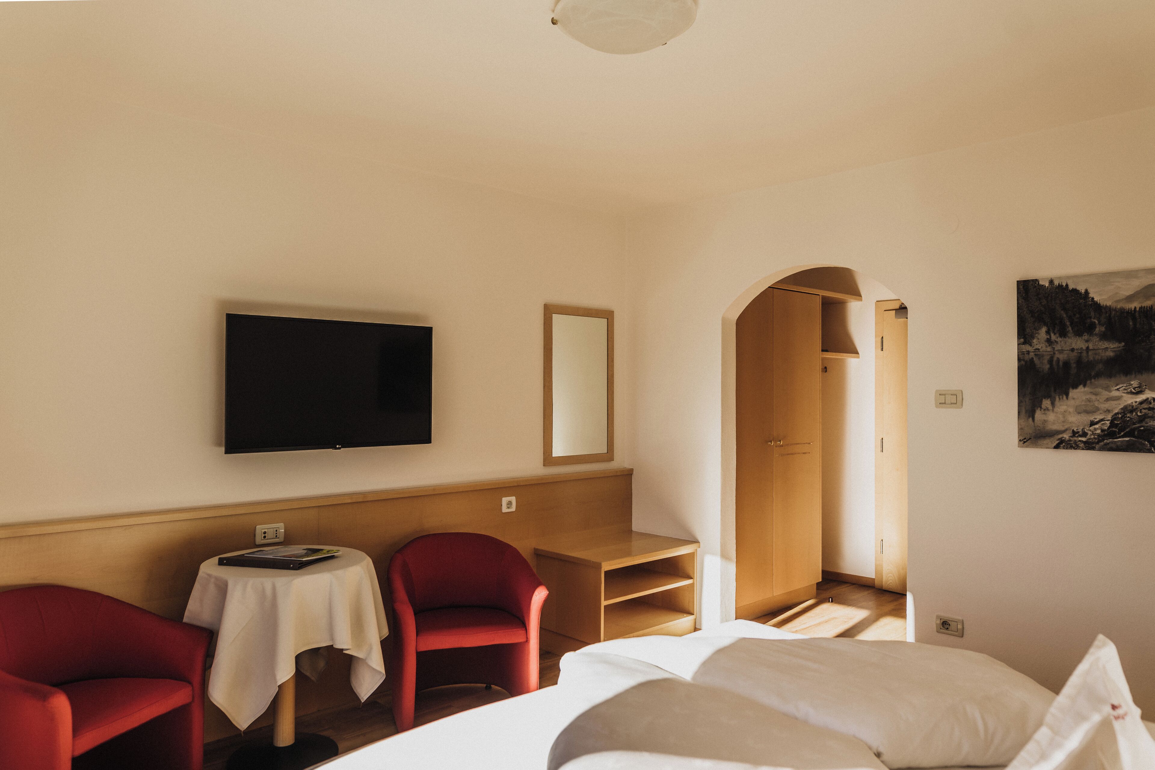 Ein Hotelzimmer mit einem Bett, einem an der Wand befestigten Fernseher, einem roten Sessel, einem kleinen Tisch mit weißer Tischdecke und einem Bild an der Wand.