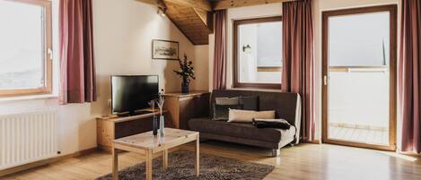 A cozy living room with a wooden ceiling, a sofa, a TV, and a large window with curtains.